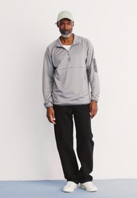 Grå halv-zip genser, lett stoff, to lommer, brukt med svarte bukser og hvite joggesko, toppet med en lys grønn cap.