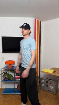 Junger Mann in einem hellblauen Hemd und schwarzer Kappe steht in einem Raum mit Fernseher, orangefarbener Lampe, blauen Regalen, Basketball und bunt gestreiftem Wanddetail.