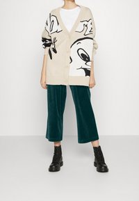 Beige gebreide cardigan met zwart cartoonfiguur ontwerp. Gecombineerd met teal corduroy wijdpijpenbroeken en zwarte veterlaarzen.