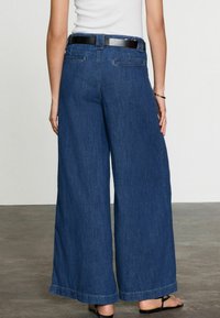 Leveled-leg denim pants vankkoissa tummansinisissä, joissa on korkea vyötärö, musta vyö ja takataskut. Lisätyönnissä sileä pinta, puhtaat saumat.