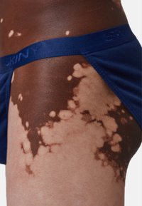 Slip di cotone blu navy con elastico testurizzato che presenta il logo "SKINY". Primo piano della gamba che mostra la pelle con tonalità e motivi vari.