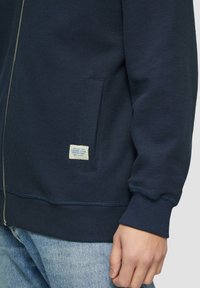 Sweatshirt zippé marine en tissu texturé, avec des poignets côtelés et une poche sur le côté. Présente une petite étiquette tissée près de la poche.