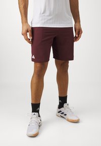 Shorts de sport bordeaux avec une texture lisse, arborant un logo Adidas blanc. Portés avec un t-shirt blanc et des chaussettes noires. Chaussures de sport blanches.