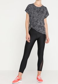 Kvinna klädd i en gråmönstrad kortärmad topp knuten i midjan, svarta leggings med nätpanel och korallfärgade träningsskor.