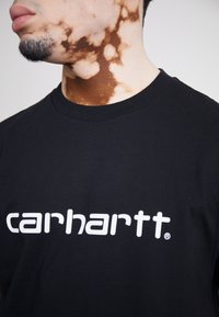 Czarny bawełniany t-shirt z białym logo "carhartt" z przodu. Zbliżenie na fakturę materiału i dekolt.
