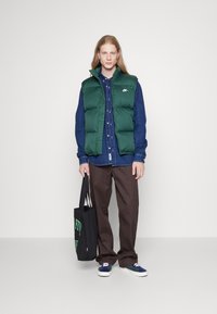 Grøn puffer vest over en blå skjorte med knapper, brune bukser og navy sneakers. Holder en sort tote bag med grøn tekst. Glatte stofoverflader.