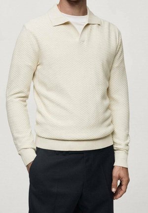 Mann trägt einen cremefarbenen, strukturierten Langarmpullover mit kleinem Kragen über einem weißen Unterhemd und dunklen Hosen.