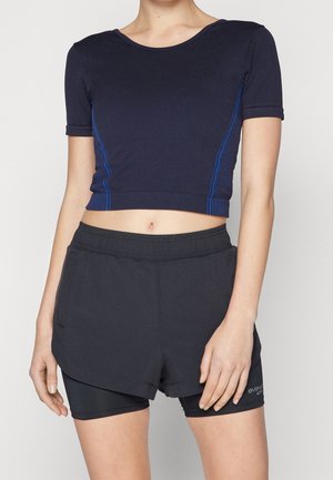 Femme portant un top court bleu marine à manches courtes avec des bandes latérales bleues et un short de sport noir à couches superposées.