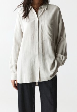 Vrouw draagt een losse, lichtbeige blouse met lange mouwen en knopen, voorzien van een borstzak, gecombineerd met een zwarte broek, staand tegen een effen achtergrond.