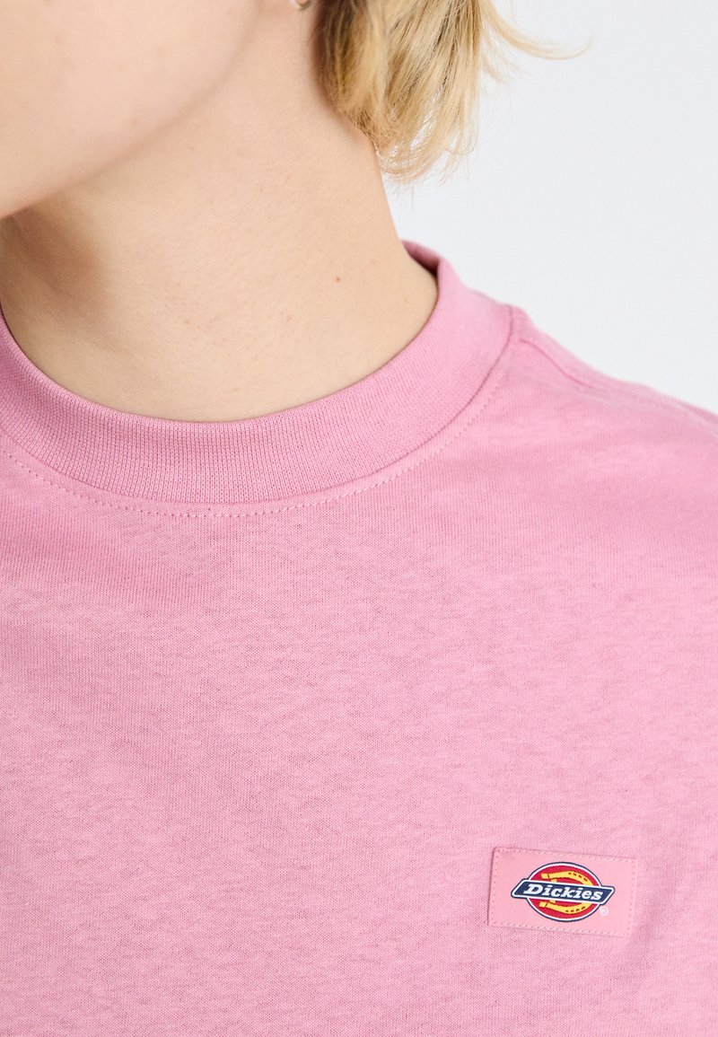 T-shirt di cotone rosa con collo rotondo, dotata di una piccola patch colorata del logo Dickies sul lato sinistro del petto. Texture liscia, design casual.