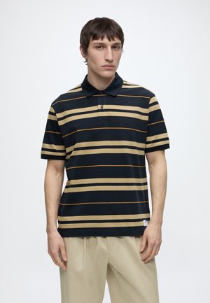 Homme portant un polo rayé noir et beige à manches courtes et un pantalon beige clair sur un fond uni.