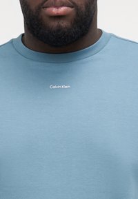 Svetlo modré bavlnené tričko s okrúhlym výstrihom, s bielym logom "Calvin Klein" umiestneným v strede hrude, hladká textúra.