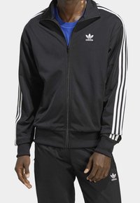 Svart atletisk jacka i smidigt tyg med vita tre-rands detaljer på ärmarna och vit adidas-logga på bröstet. Full dragkedja.