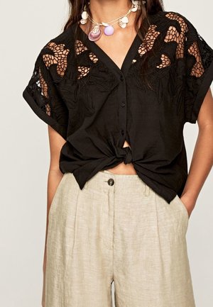 Zwart kort mouwen overhemd met knopen en bloemrijke kant details, knoop gedragen over beige linnen broek met hoge taille en een schelpenketting.