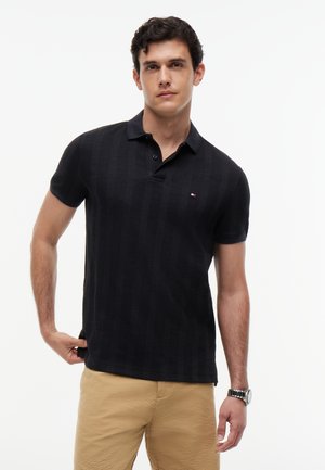 Mand iført sort kortærmet poloshirt med lille logo på brystet og beige teksturerede bukser, poserer mod hvid baggrund.