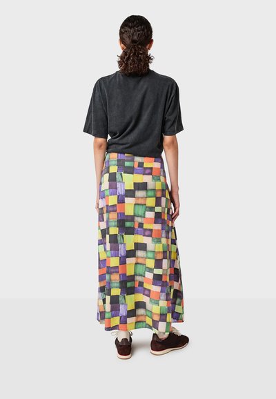 Personne avec des cheveux bouclés en queue de cheval, portant une chemise sombre à manches courtes, une jupe en patchwork coloré, des chaussettes blanches et des baskets sombres, de dos.