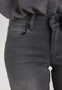 Los pantalones vaqueros de denim gris presentan un diseño ajustado, estilo de cinco bolsillos y una textura sutil con un patrón de sarga visible. Cierre clásico con botón y cremallera.