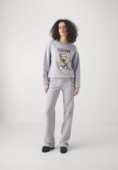 Szürke kapucnis pulóver és nadrág szett, amelyen egy teddy maci grafika és "Moschino" felirat található. Puha anyagból készült, lazán illeszkedik és bordás mandzsettával rendelkezik.