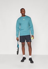 Sudadera azul claro con un pequeño logo verde, combinada con pantalones cortos negros, sosteniendo una raqueta de tenis y una pelota. Zapatillas negras y calcetines blancos.