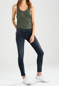 Olivgrön linne i kombination med åtsittande mörkblå jeans och vita sneakers. Outfiten har en avslappnad design och en smal silhuett.