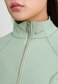 Gaiši zaļš ar rāvējslēdzēju augstākā apkakle, ar sudraba rāvējslēdzēju un Nike swoosh logotipu. Gluda tekstūra un pieguļošs dizains ar šuvju detaļām.