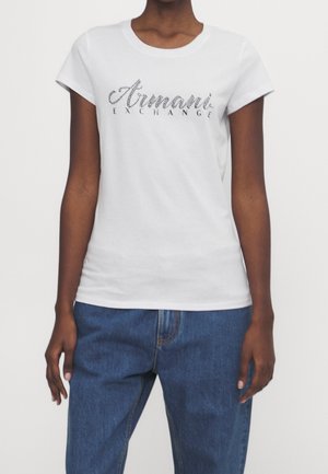 Osoba nosząca białą dopasowaną koszulkę z logo "Armani Exchange" i niebieskie dżinsy, pokazana od ramion do bioder.