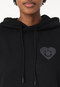 Black hoodie with ribbed texture, featuring a heart design in dark gray with the letters "HGO" and a smiling face. Drawstrings are present.