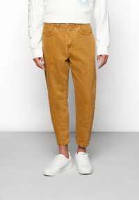 Pantaloni din catifea maro, cu picioare conice, buzunare frontale și detalii vizibile ale cusăturii, asortați cu adidași albi.