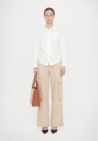 Camicia bianca con taschino, pantaloni cargo beige con tasche laterali, borsa marrone con dettagli borchiati e scarpe nere a punta.