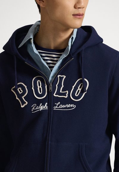 Moški, oblečen v temno modro jopa z kapuco in zadrgo, na katerem je izvezen napis "POLO Ralph Lauren", oblečen prek svetlo modre srajce z ovratnikom in temno modre majice z črtami.