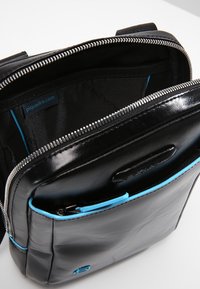 Borsa in pelle nera con chiusura zip, caratterizzata da una texture liscia, dettagli blu e una tasca interna. L'etichetta del marchio è visibile all'interno.