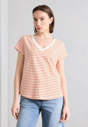 Nuori nainen oranssia ja valkoista raidallista V-kaulaista T-paitaa ja vaaleansiniset farkut, seisoo yksinkertaista vaaleanharmaata taustaa vasten.
