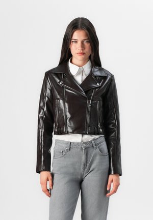 En ung kvinne står i en kort, glanset sort skinnjakke over en hvit skjorte, kombinert med grå jeans, mot en enkel hvit bakgrunn.