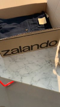 Tamsiai mėlyni džinsai, sulankstyti Zalando prekės ženklo kartoniniame dėžutėje, padėti ant marmurinio stalviršio, su kainos etikete, matoma ant audinio.