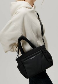 Svart quiltad handväska med dubbla handtag och en avtagbar axelrem. Har en minimalistisk logotyp, mjukt tyg och en strukturerad form.