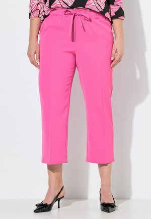 Kobieta ubrana w jaskrawo różowe, krótkie spodnie z troczkiem, czarne buty na obcasie z paskiem z tyłu oraz różowo-czarną wzorzystą bluzkę.