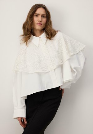 Hajában hullámos barna hajú nő, fehér blúzt csipkeköpenyével és fekete nadrágot visel, egy sima világos háttér előtt áll.