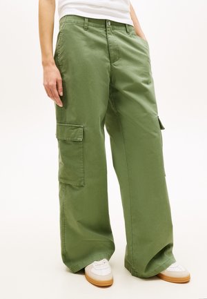 Pantaloni cargo largi de culoare verde măsliniu cu buzunare laterale, purtați cu adidași albi și o cămașă albă băgată în pantaloni.