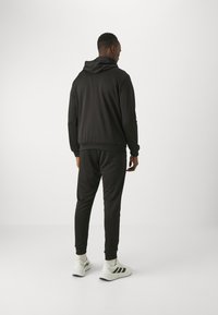 Czarna bluza z kapturem i kieszenią kangurką, w zestawie z pasującymi dresami. Posiada ściągacze na rękawach i gładką fakturę. Model nosi białe trampki.