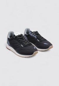 Zapatillas deportivas en negro y gris con una parte superior de tela texturizada, diseño de cordones, suela de color claro y un sutil logo en la lengüeta.