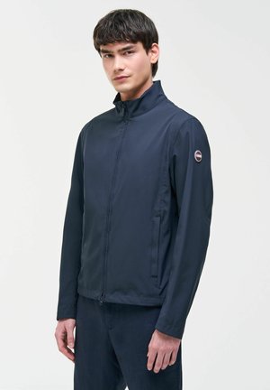 Ung mand iført en mørkeblå lynlåstrøje med høj krave og logo-patch på venstre ærme, stående mod en ensfarvet lys baggrund.