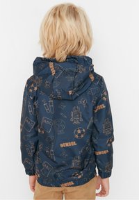 Child with blonde hair wearing a navy blue hooded jacket featuring school-themed icons, standing facing away against a plain white background.