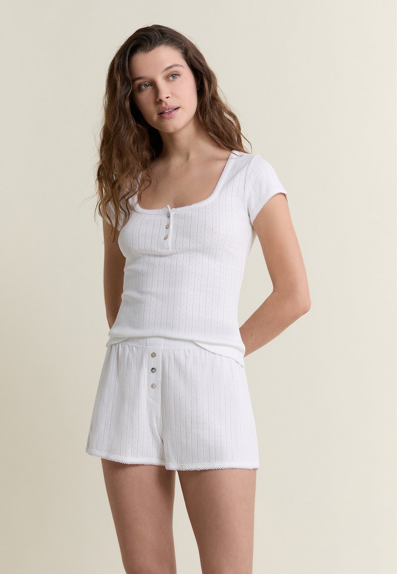 Giovane donna con capelli lunghi e ondulati che indossa una maglietta a maniche corte a coste bianca e pantaloncini bianchi coordinati, in piedi davanti a uno sfondo uniforme.