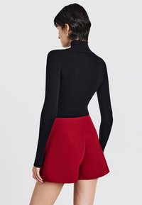 Donna con i capelli corti e scuri che indossa un maglione a collo alto nero a coste e pantaloncini rossi a vita alta su misura, rivolta di spalle contro uno sfondo bianco semplice.