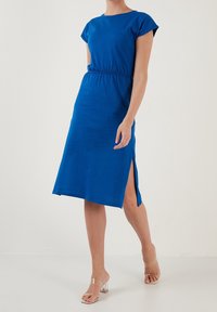 Kvinna i en blå midiklänning med korta ärmar och en sidoöppning, i kombination med sandaler med klara remmar och blockklackar, ståendes mot en enfärgad bakgrund.