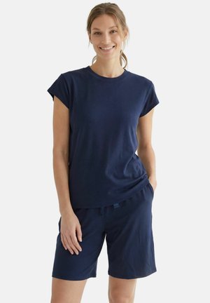 Vrouw die glimlacht, draagt een marineblauw T-shirt met korte mouwen en bijpassende knielange korte broek, staat met één hand in de zak.