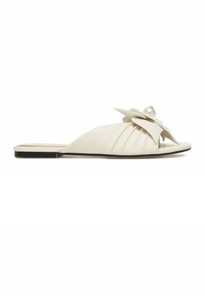 Flache beige Slip-On-Sandale mit plissiertem Obermaterial, verziert mit einer großen blumenförmigen Schleife und einem einzelnen Perlenakzent oben.