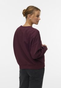 Vrouw met blond haar in een knot, gekleed in een bordeauxrode sweatshirt en zwarte jeans, zijwaarts kijkend tegen een witte achtergrond.