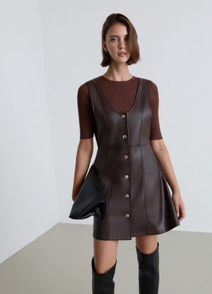 Donna che indossa un vestito marrone in pelle con bottoni sopra una maglia marrone a coste, tiene una pochette nera e indossa stivali neri al ginocchio.