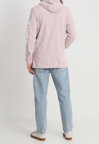 Růžová mikina s kapucí, zdobená bílými grafikami na rukávech. Nosená s světle modrými džíny a bílými teniskami. Hladká textilie.